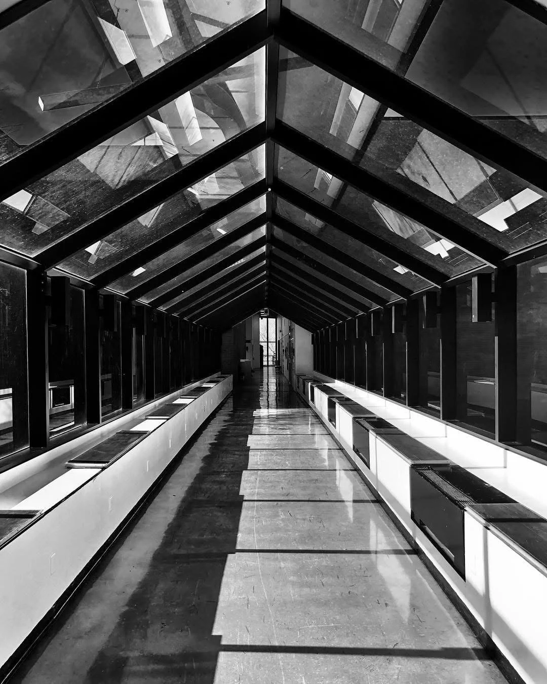Black and white photograph of a modern, enclosed walkway with glass walls and ceiling. The walkway has a polished floor with reflections and white benches along both sides. The ceiling features a series of angled glass panels that create a geometric 