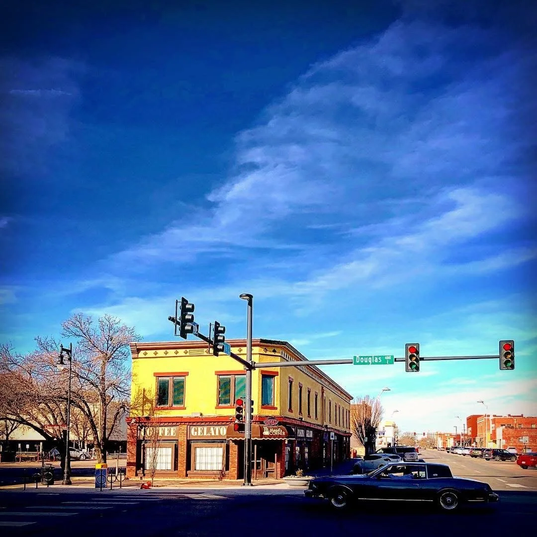 A street corner with a yellow two-story building with a gelato shop, cars parked on the street, and traffic lights, on a sunny day with a blue sky.