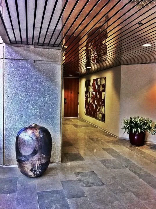 Lobby with a large decorative vase on the floor, an abstract wall art piece, potted plant, and a wooden door, with a reflective ceiling and tiled floor.