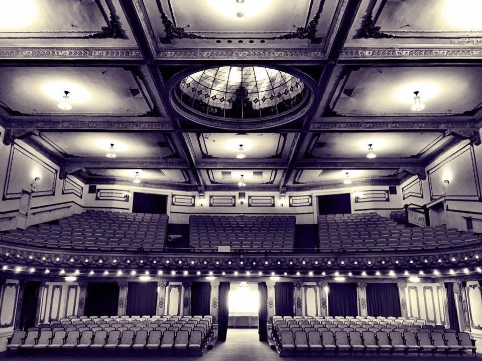 An empty historic theater with ornate interior details, a large stage, and tiered seating.