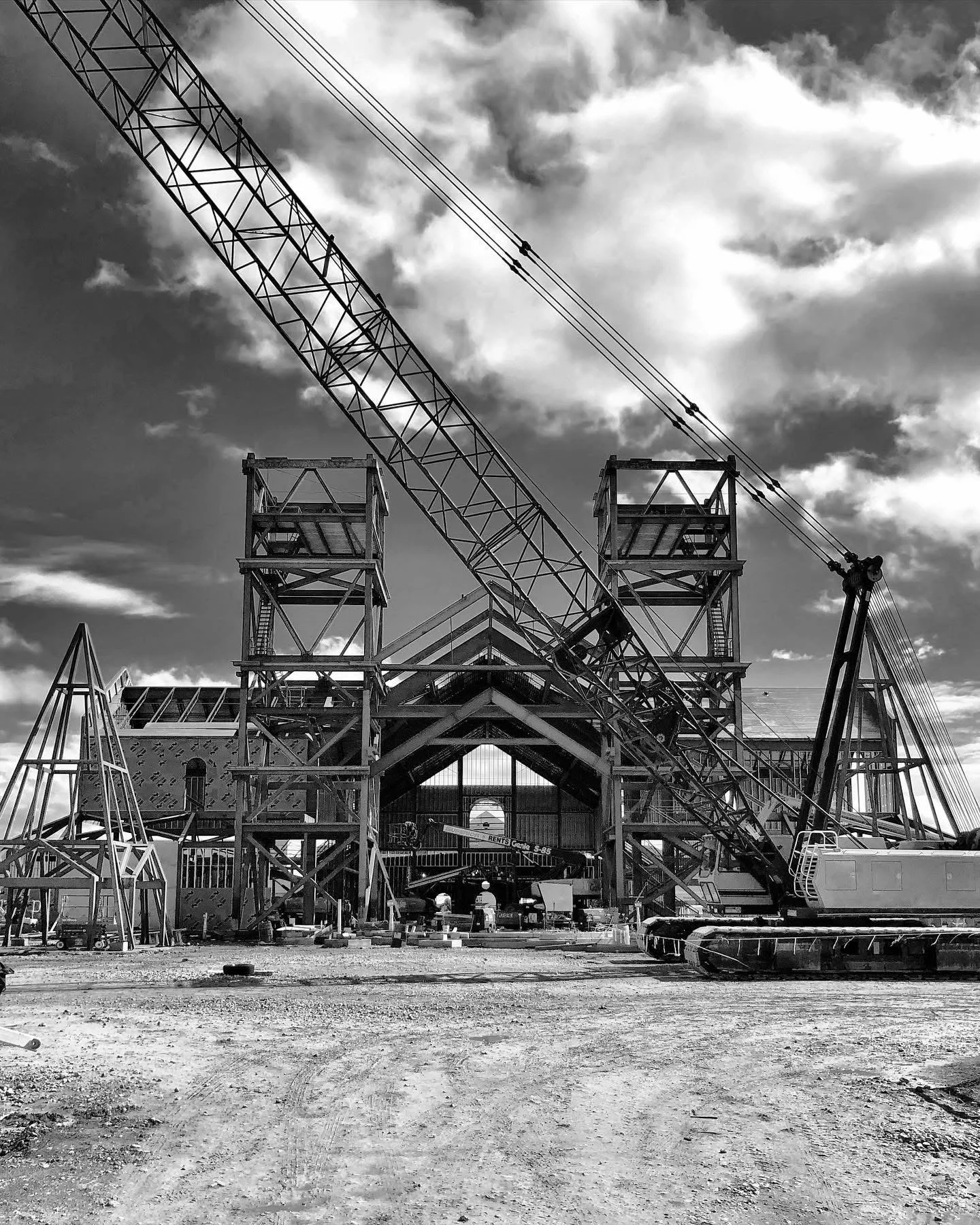 Black and white photo of a construction site with a large crane, scaffolding, and partially built structure under a cloudy sky.