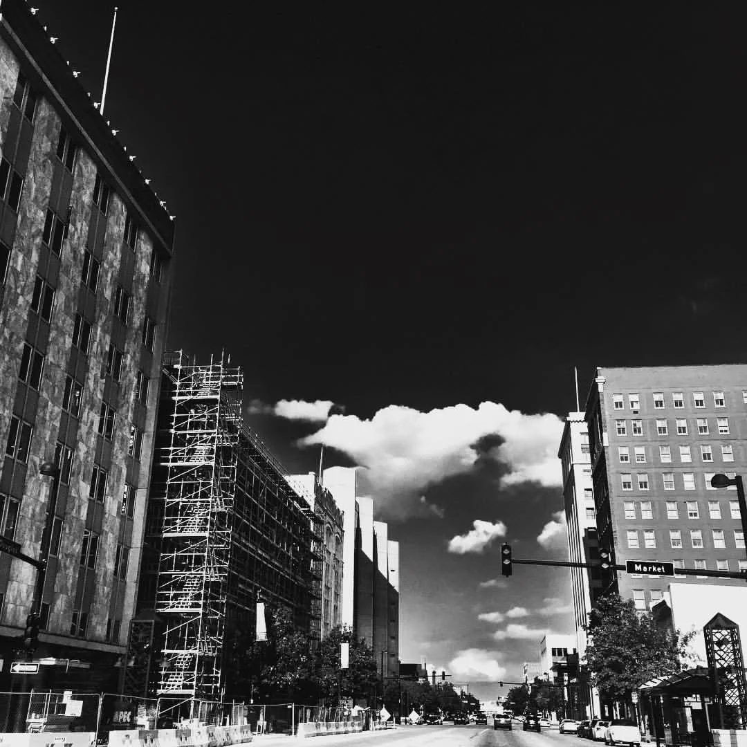 Black and white photo of city street with buildings, trees, traffic lights, and cars, under a mostly cloudy sky.