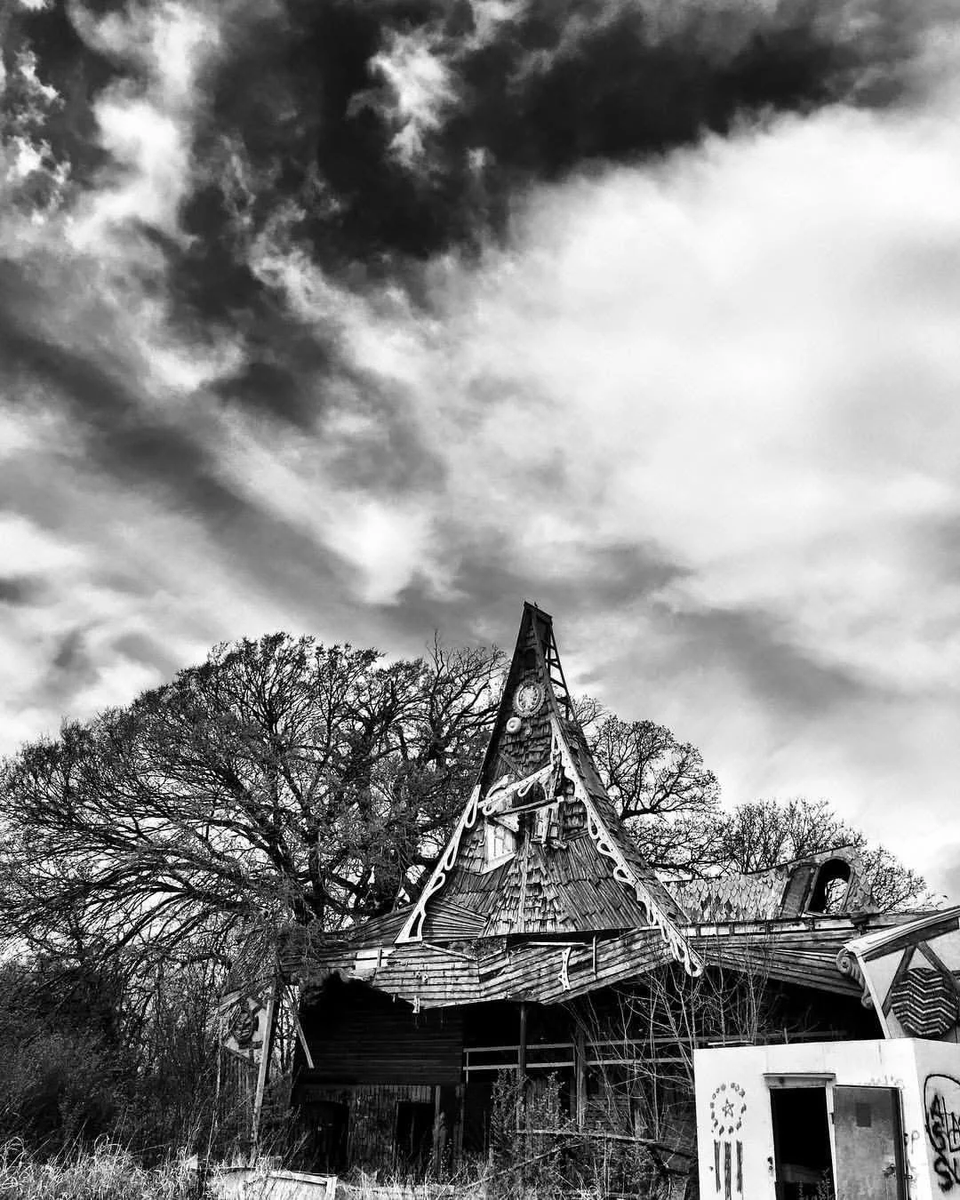 An abandoned wooden house with a steep, pointed roof, surrounded by leafless trees under a cloudy sky.