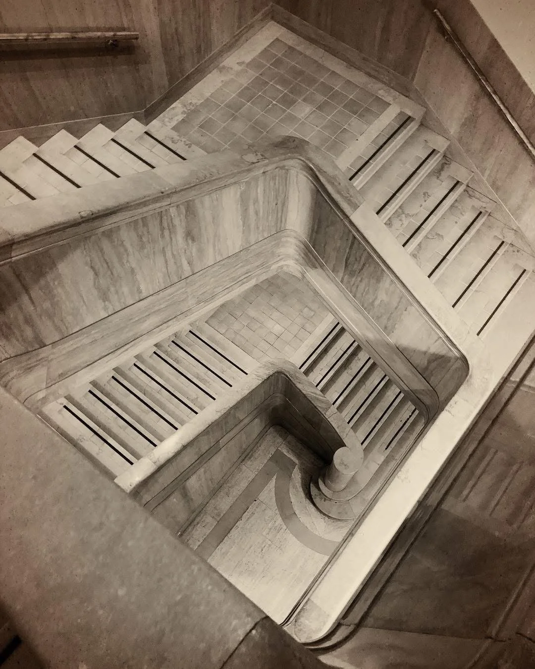 A top-down view of a marble staircase with multiple landings and dark wood railings, leading down through a building.