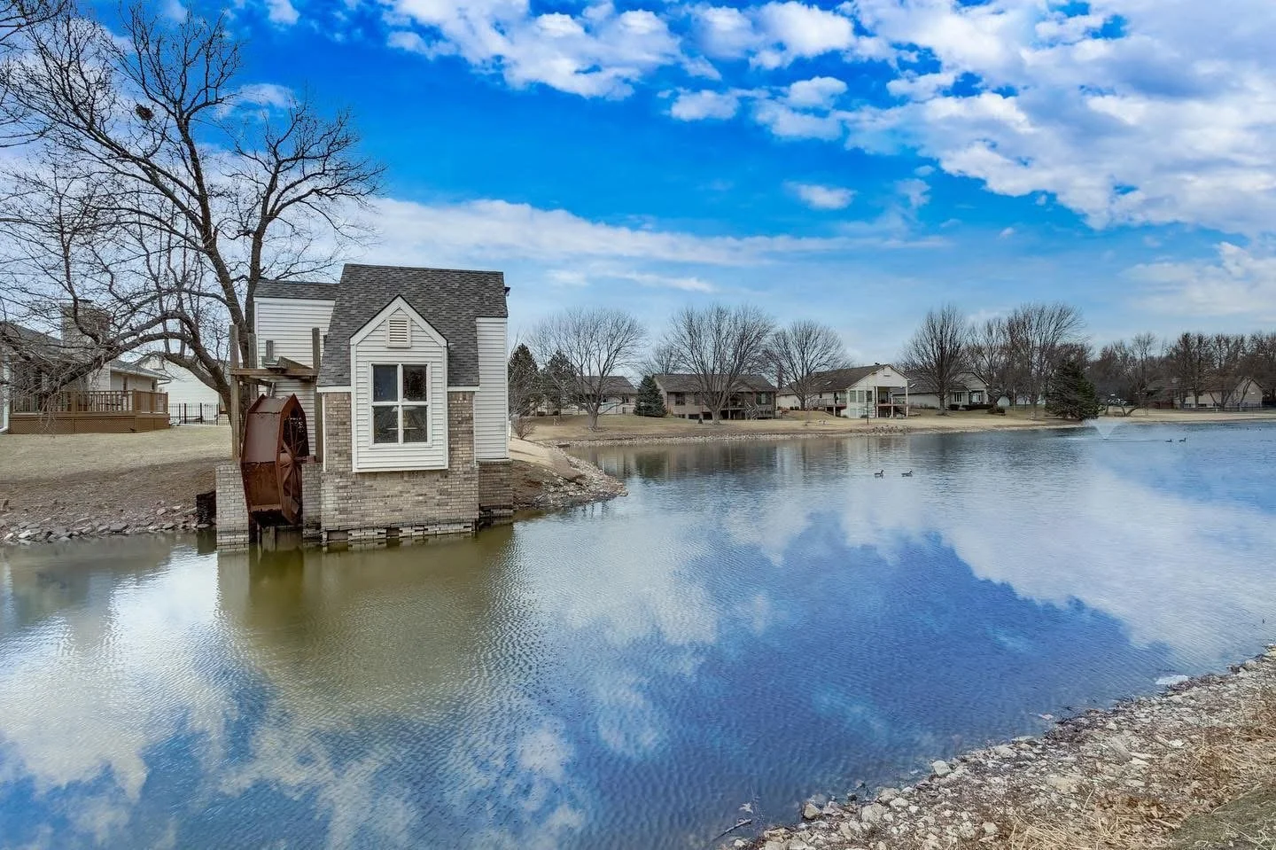 A lakeside scene with a small watermill house on the left, surrounded by leafless trees and neighboring houses across the lake; blue sky with some clouds and waterfowl in the lake.