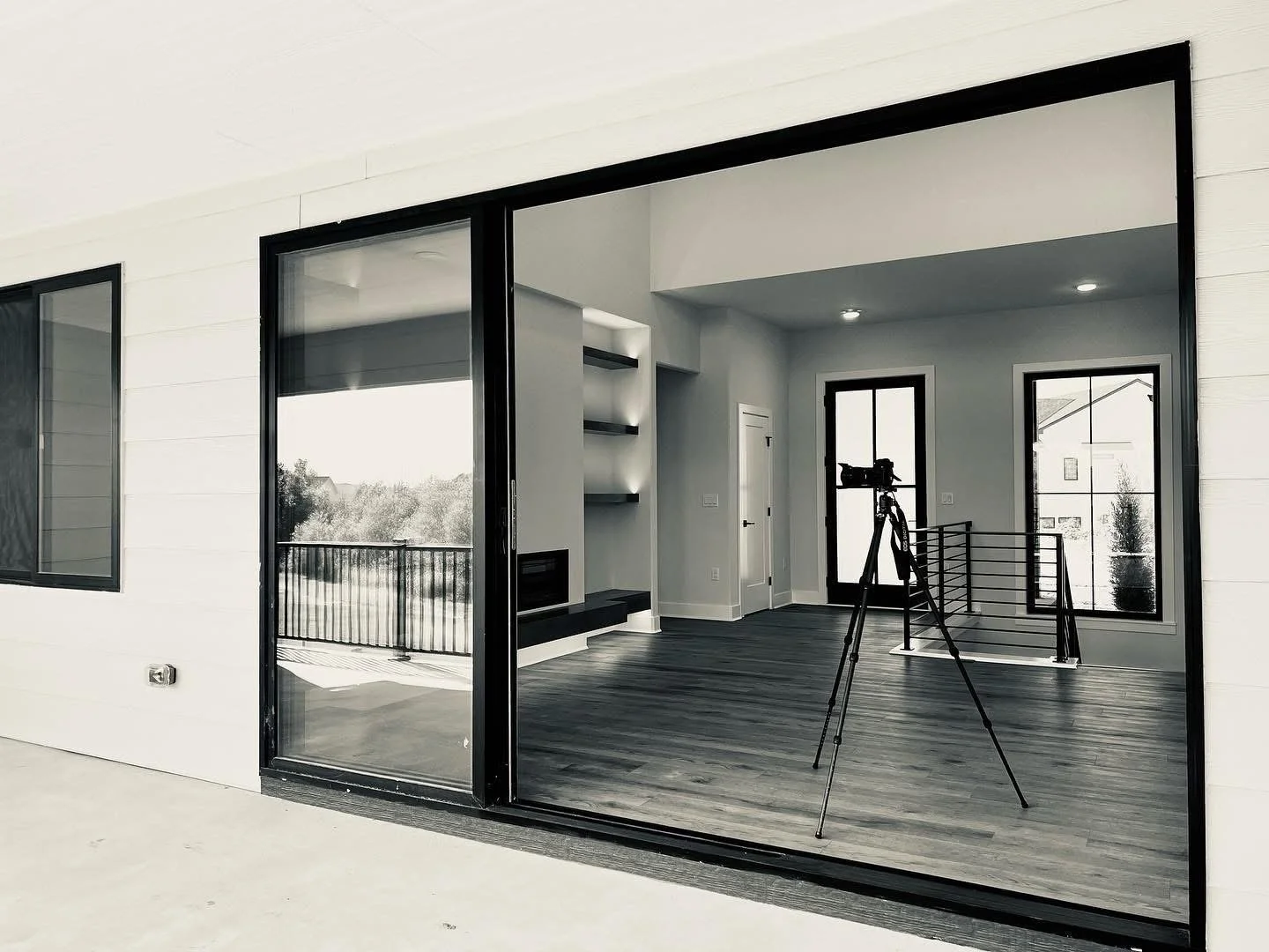 Interior view of a modern, minimalistic room with hardwood floors, large windows, and a sliding glass door opening to a balcony with a black railing. There is a camera on a tripod facing the room, and a fireplace with shelves nearby.