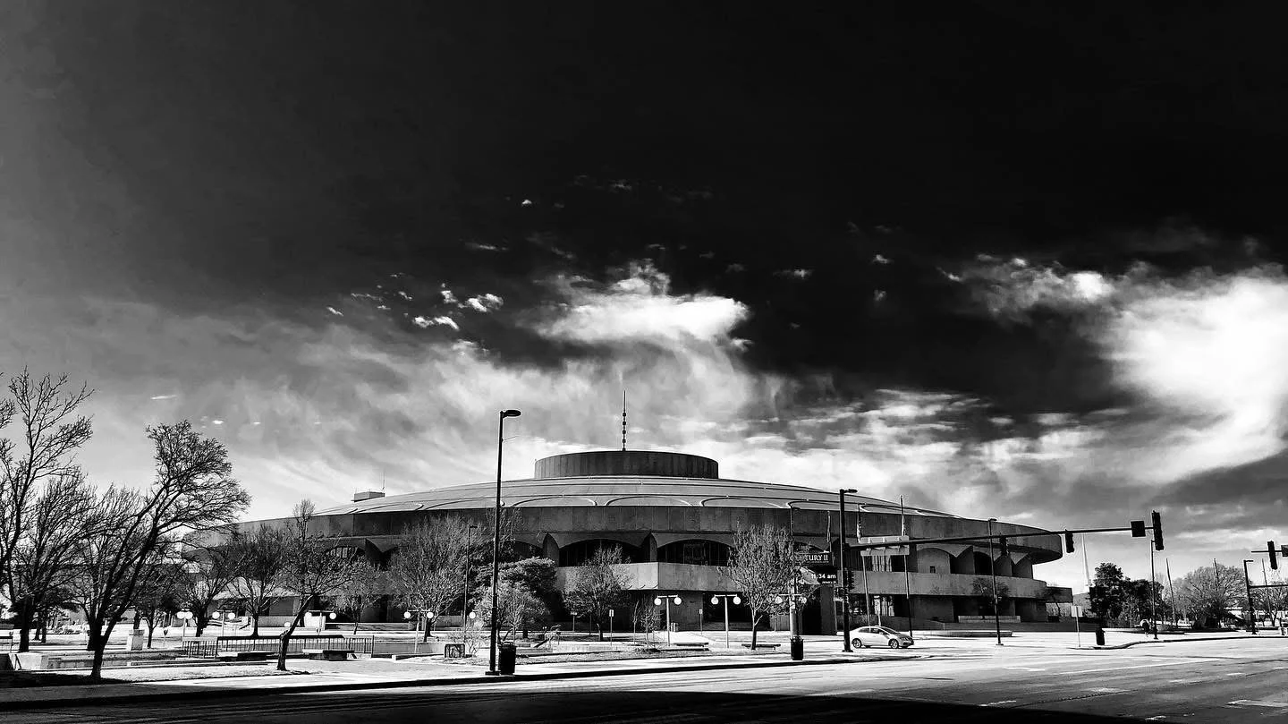 Black and white photo of a circular auditorium or concert hall with a futuristic design, surrounded by streetlights, trees, and traffic signals under a cloudy sky.