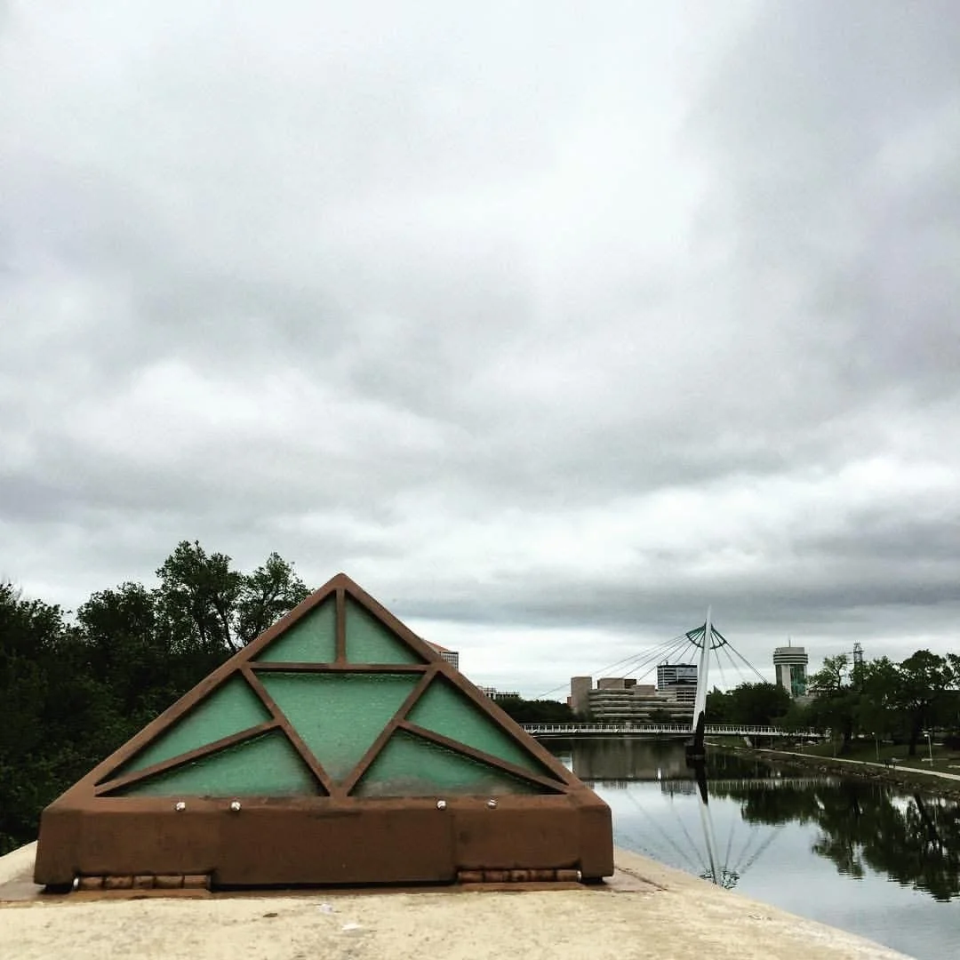 A rooftop view featuring a triangular rooftop vent with green glass panels, a canal or river, a modern pedestrian bridge, and city buildings under a cloudy sky.