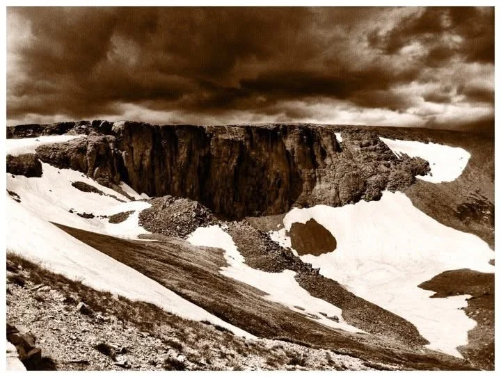 Mountain landscape with snow patches, rocky slopes, and a dramatic cloudy sky.