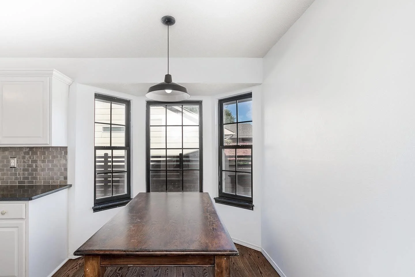 Dining area with three large windows, a wooden table, and a black pendant light hanging from the ceiling.