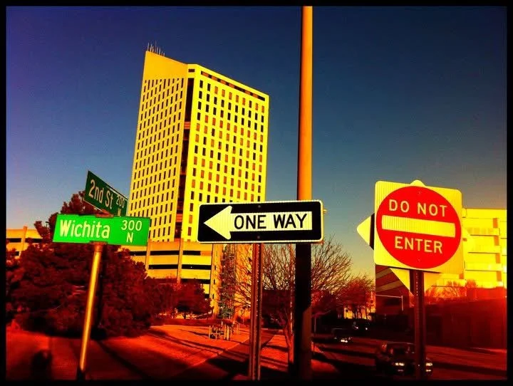 Street intersection with Bingo height department building, green street sign for Wichita 300 N, black and white one-way sign pointing left, and red and white 'Do Not Enter' sign, with clear sky and trees.