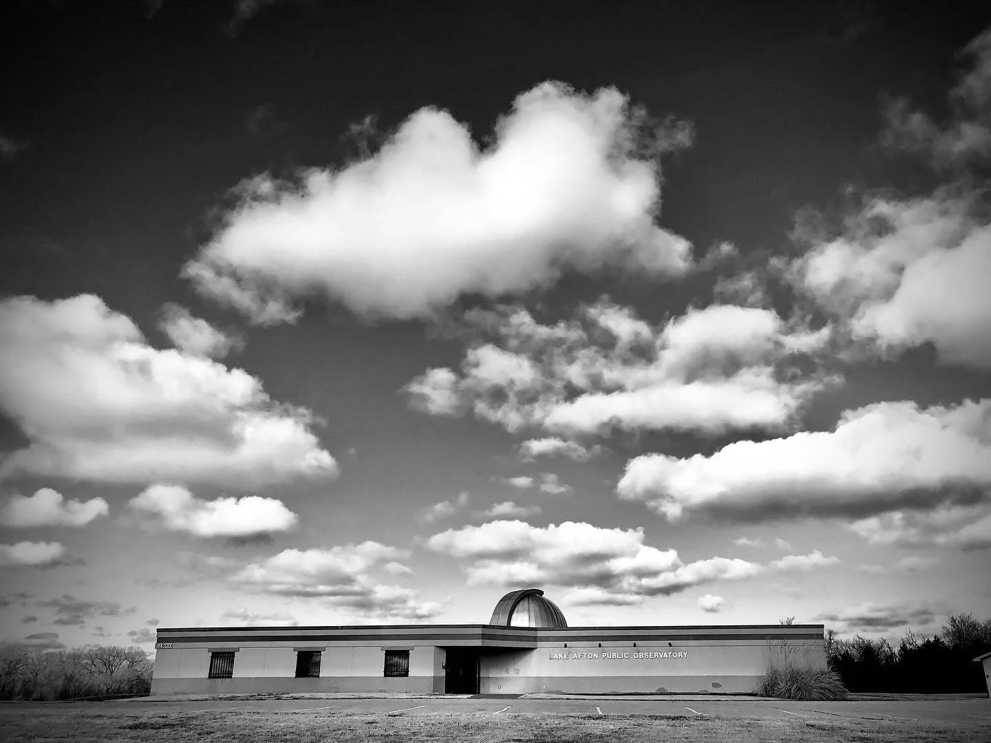 Black and white photo of Lake Afton Public Observatory building with a domed roof, three visible windows, and an entrance door, under a partly cloudy sky.
