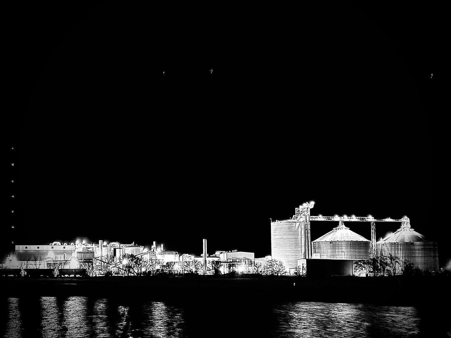 Nighttime industrial landscape of factory buildings with silos, some emitting smoke, reflecting on a body of water in the foreground.