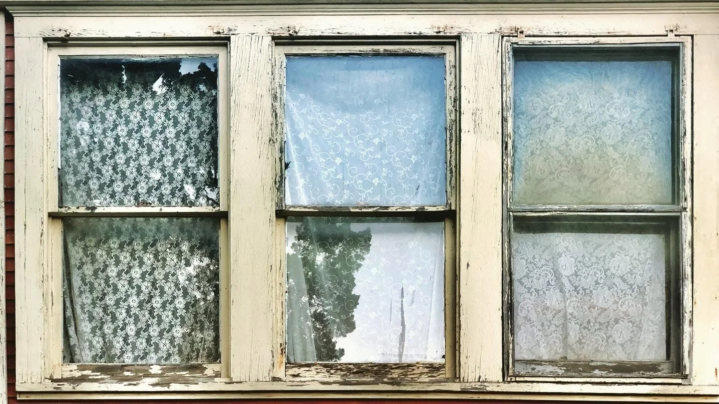 Four-pane window with peeling white paint and lace curtains, surrounding a reflection of trees and sky.