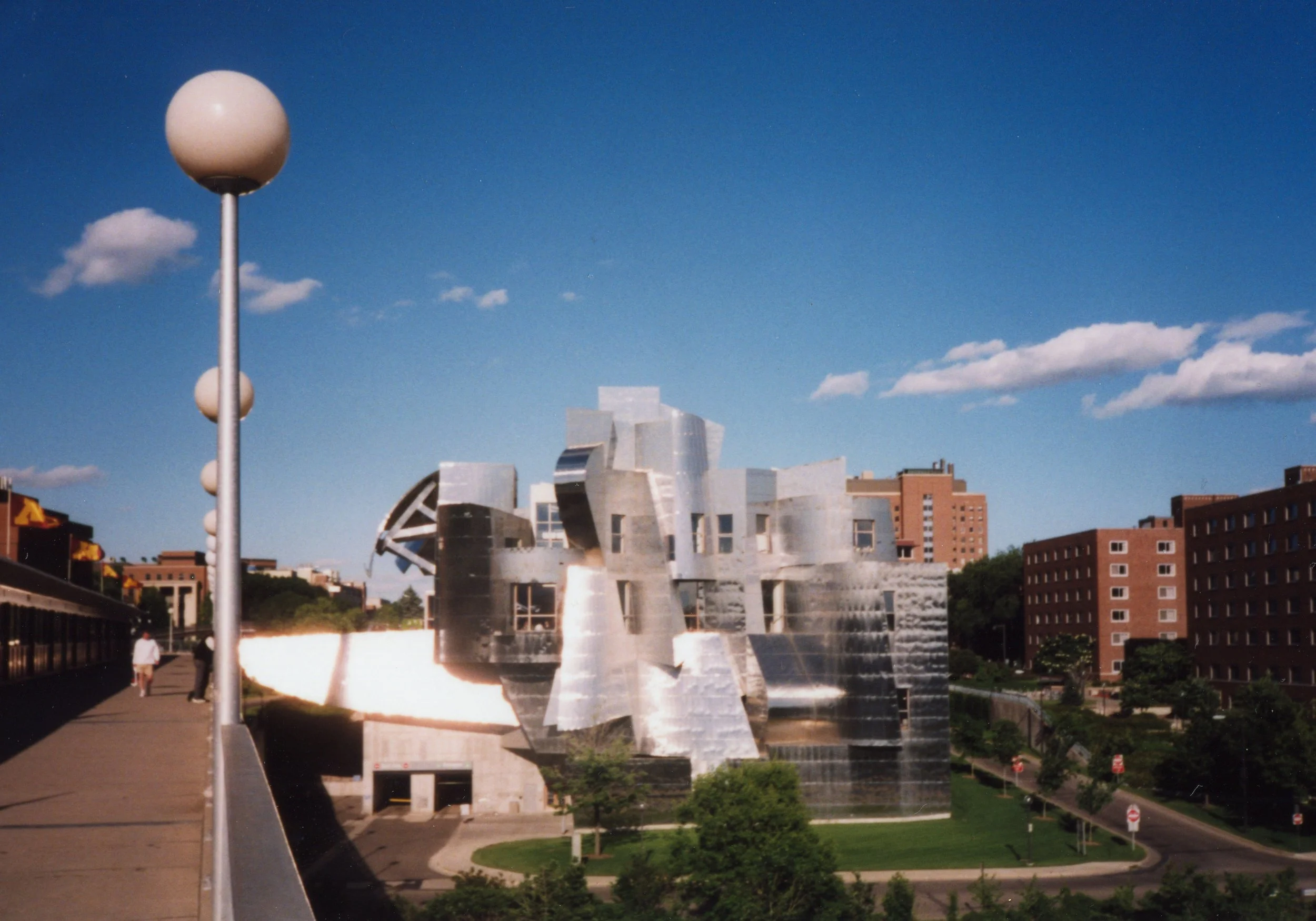 Reflective metallic modern building with irregular shapes under a blue sky with scattered clouds, located in an urban park with trees and pathways.