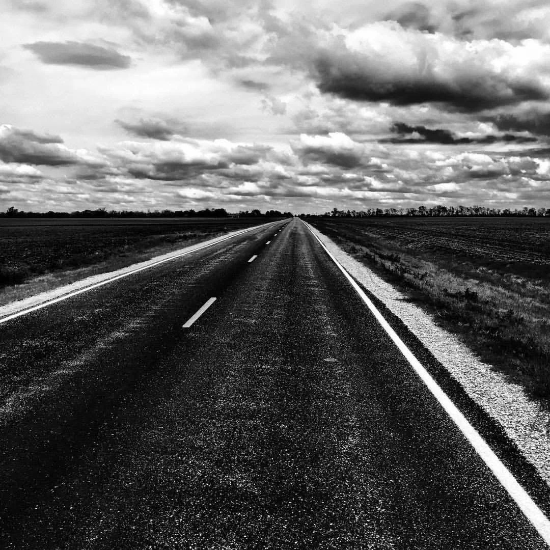 A long, straight rural highway stretching into the horizon under a cloudy sky, in black and white.