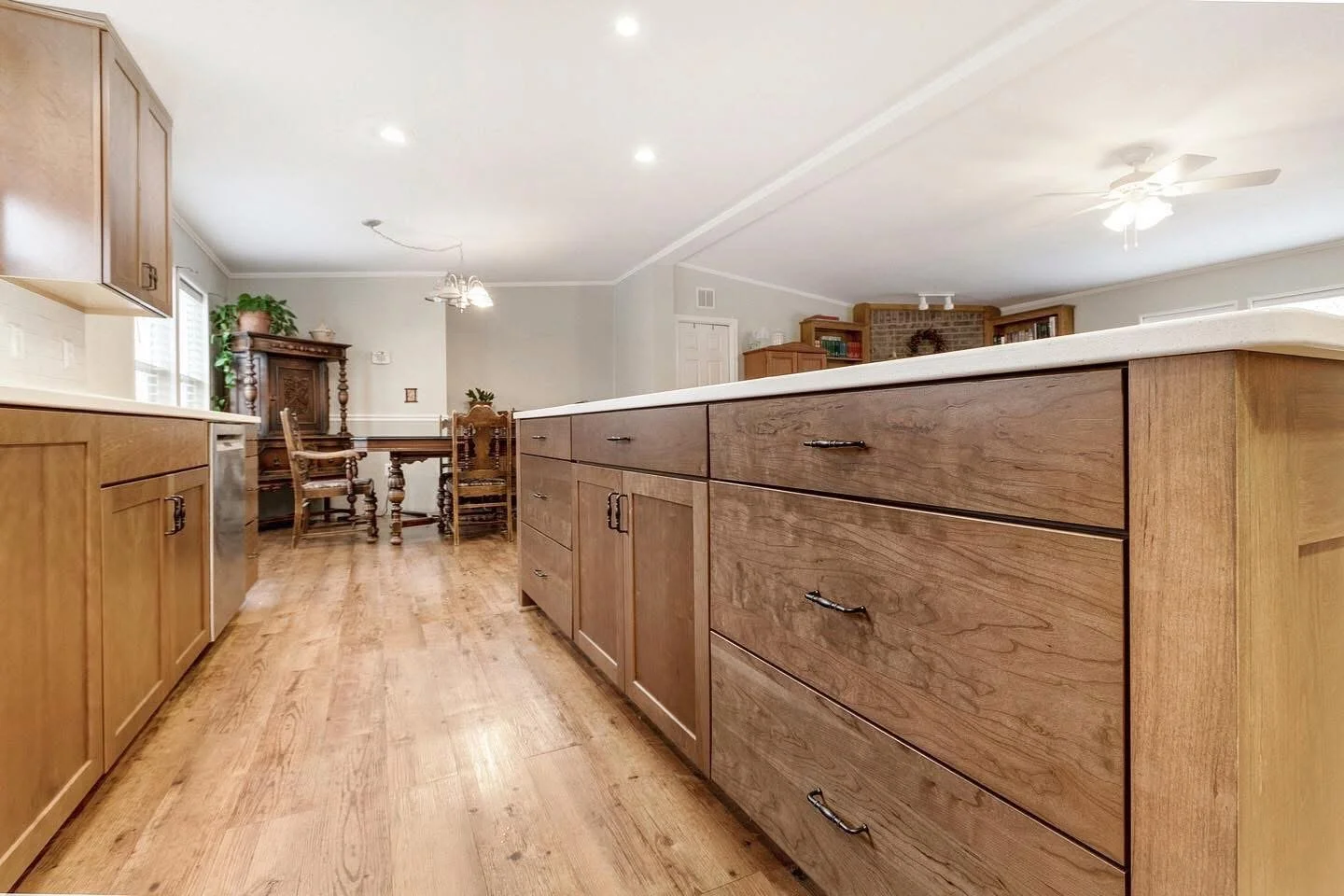 View of a spacious kitchen with wooden cabinets, a long central island, hardwood floors, and a dining area with a table and chairs in the background.