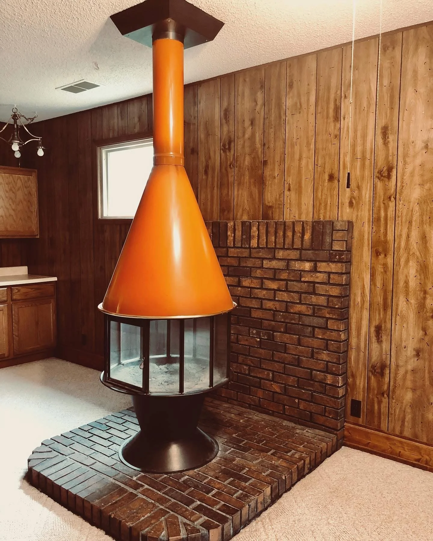 A vintage floating fireplace with an orange cone-shaped hood, brick base, and glass enclosure in a wood-paneled room.