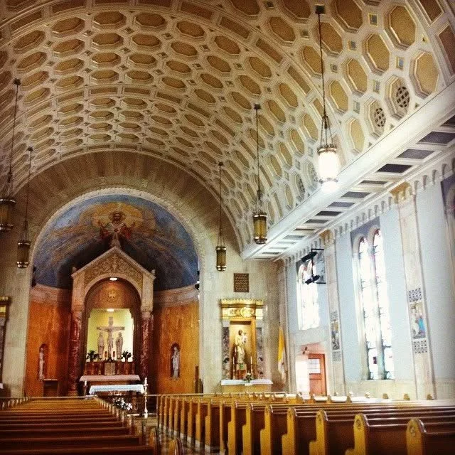 Interior of a church with wooden pews, arched ceiling, stained glass windows, and religious statues.