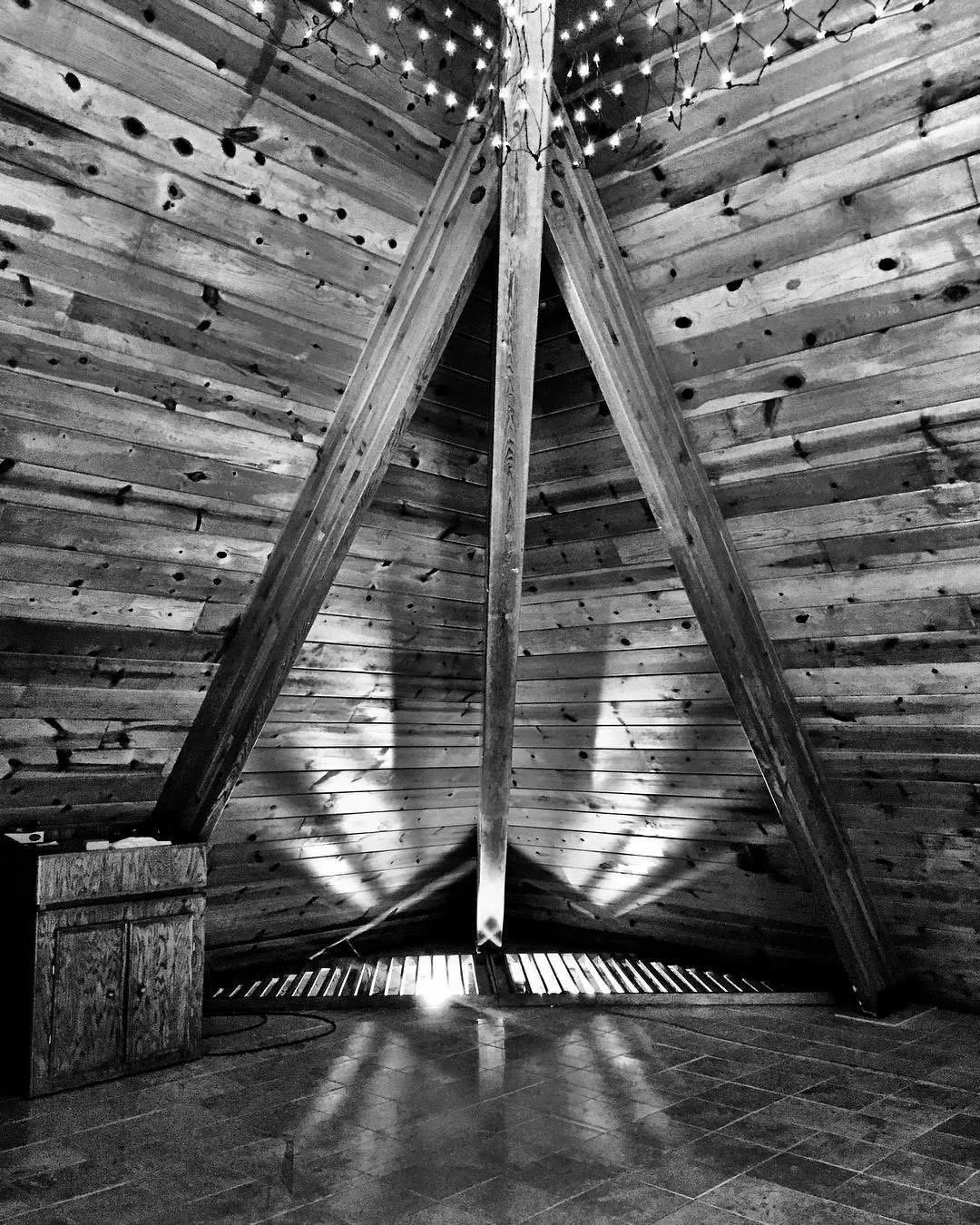 Black and white photo of an attic with wooden ceiling and walls, illuminated by string lights hanging from the apex. Light shines through gaps near the bottom, creating a pattern of shadows on the tiled floor.