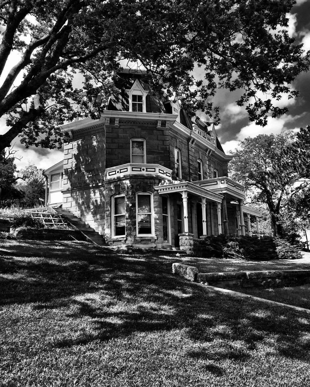 A large, historic Victorian-style house with ornate architectural details, tall windows, and a stone facade, surrounded by trees and a well-maintained lawn in black and white.