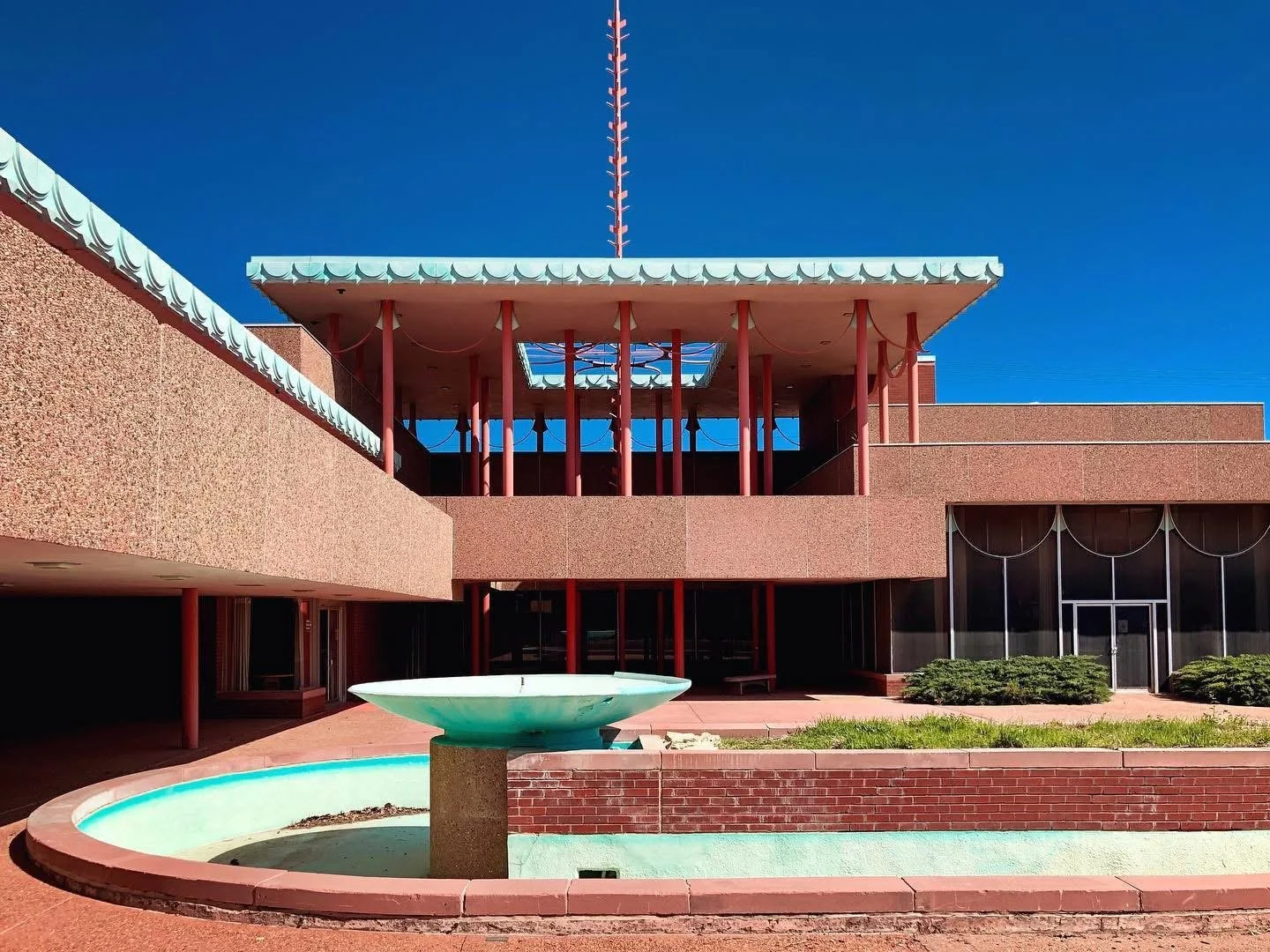 Mid-century modern building with red columns, a decorative fountain in front, and a blue sky.