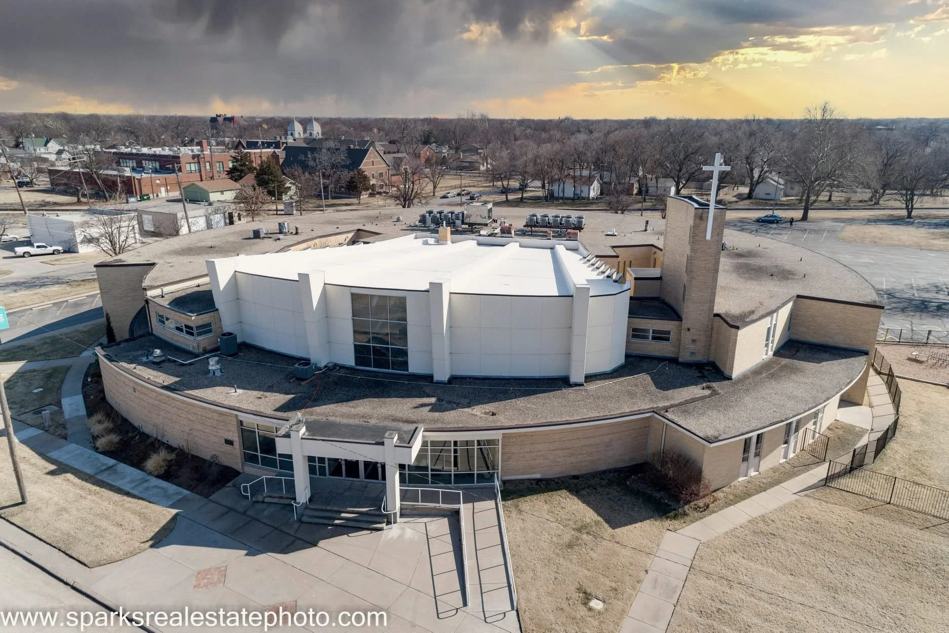 Aerial view of a modern church building with a cross on a brick tower, surrounded by a parking lot and sidewalks in a suburban area, with houses and trees in the background under a cloudy sky.