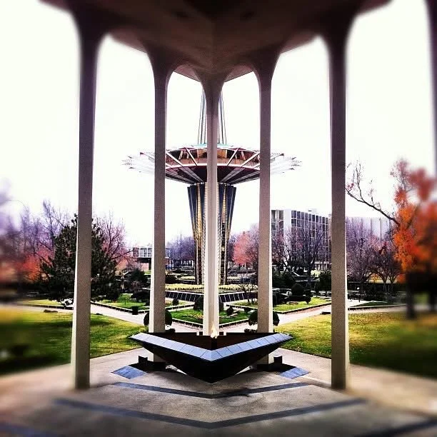 Symmetrical view of a modern outdoor pavilion with a futuristic structure in the park, featuring tall supporting columns and a platform with benches, surrounded by trees and city buildings in the background.