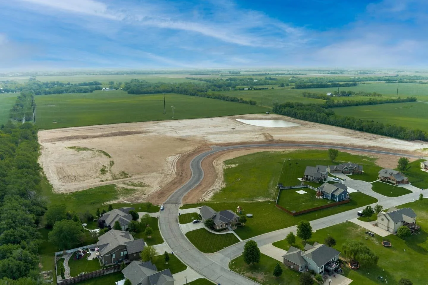 Aerial view of a suburban neighborhood with houses, green lawns, and a construction site in the background with bare soil and a small pond.