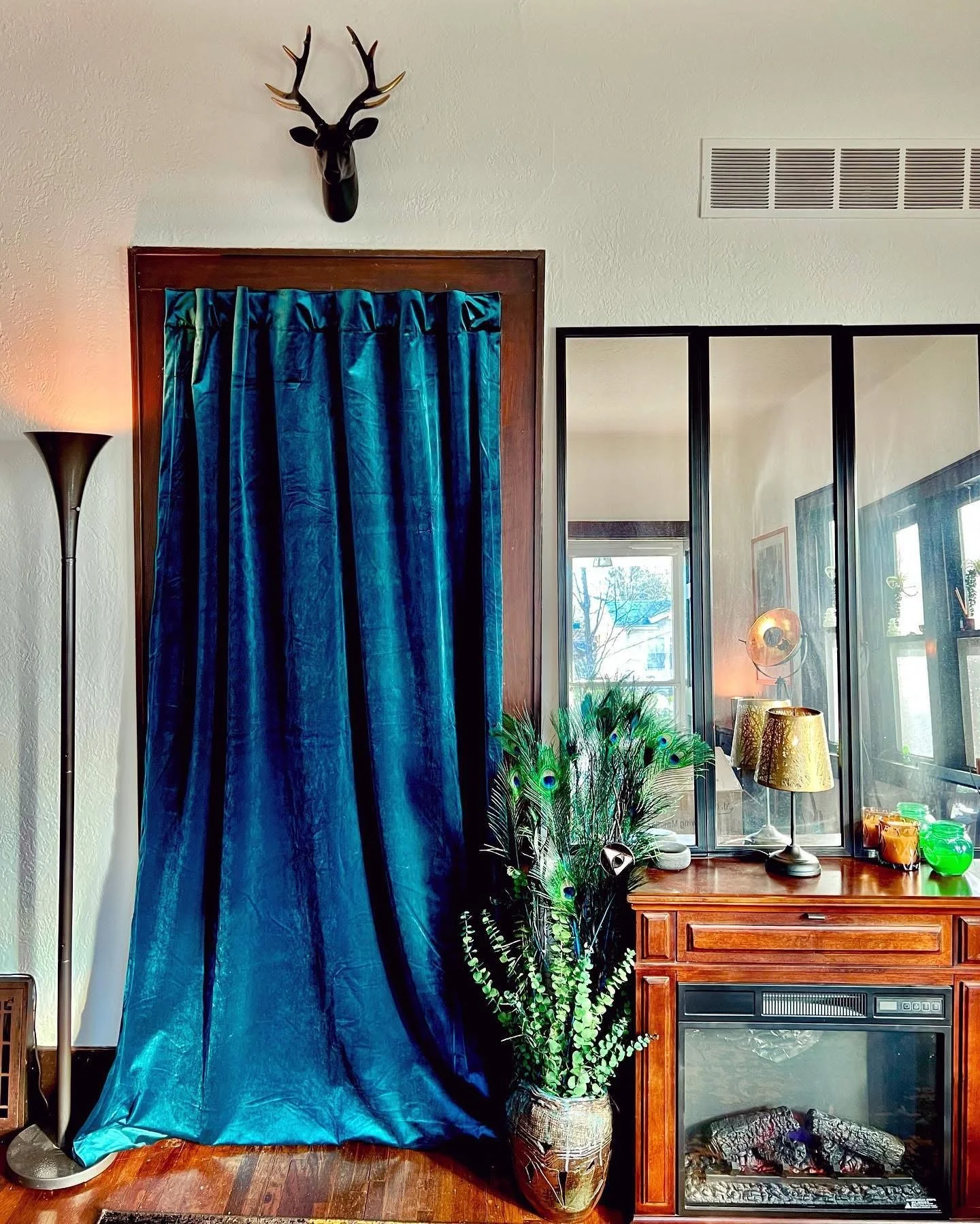 A living room corner with a dark snake-wood frame blue velvet curtain, a potted peacock feather plant, a wood cabinet with a glass fireplace, gold table lamps, and a mirror reflecting a window and other decor.
