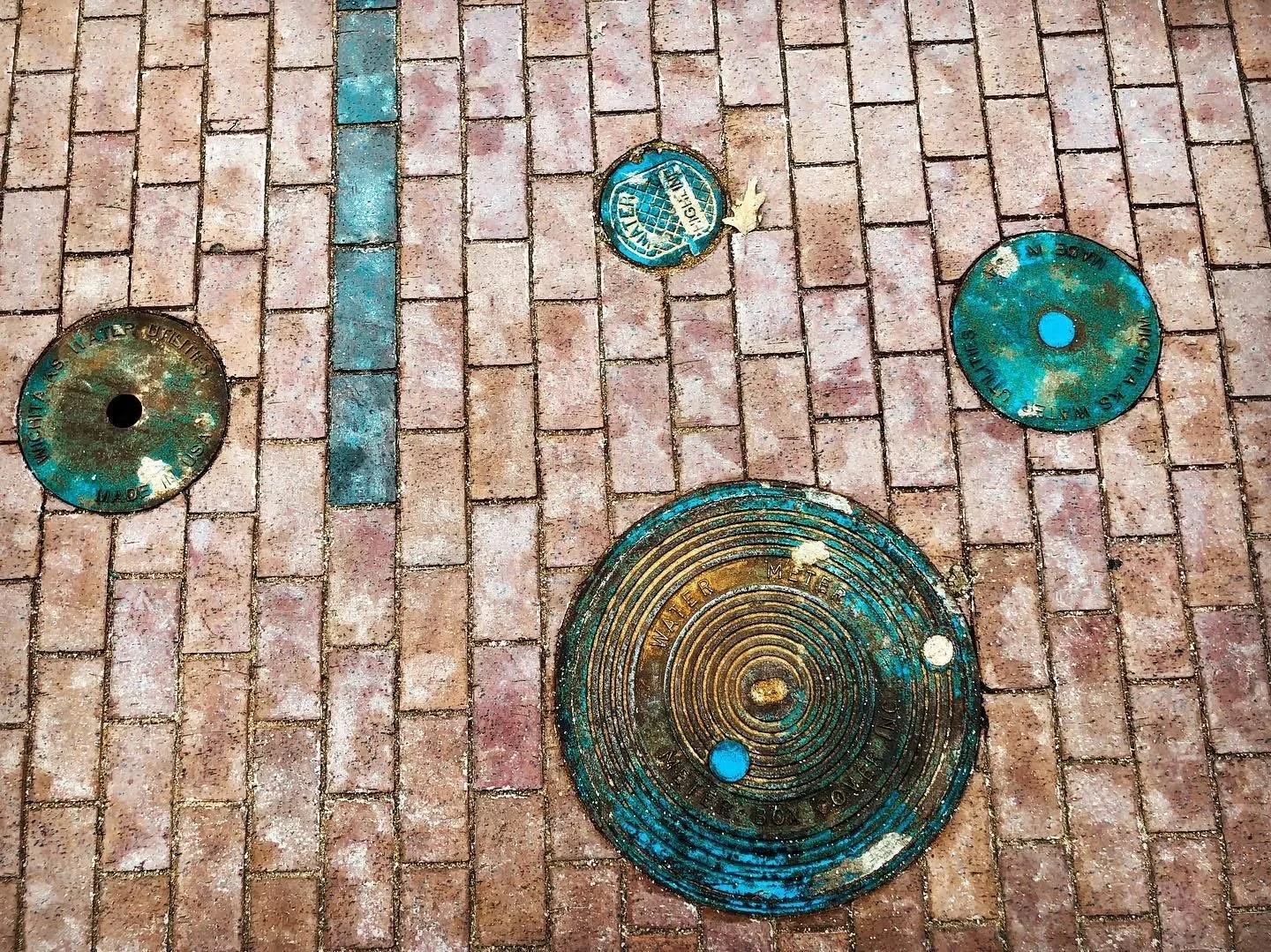 Four circular utility access covers on red brick sidewalk, with blue-green patina and one with concentric circles, one with a peace symbol sticker.