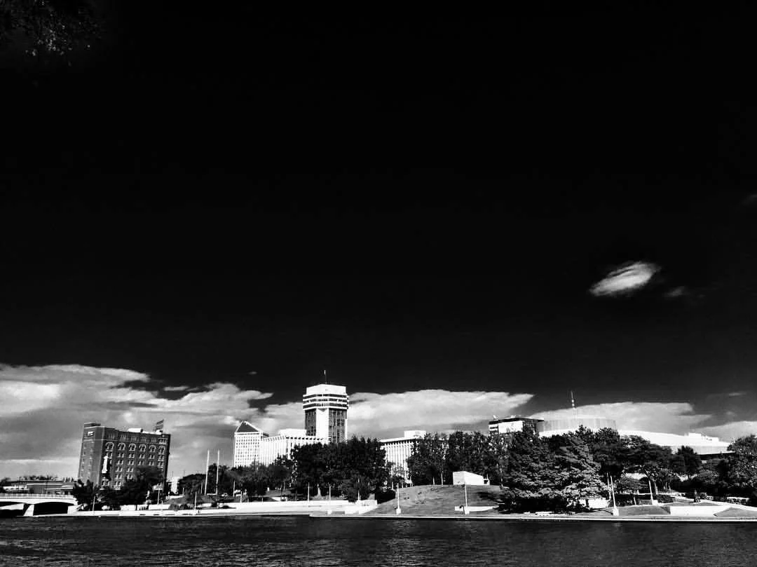 Black and white photo of a city skyline with tall buildings, trees, and a body of water in the foreground.