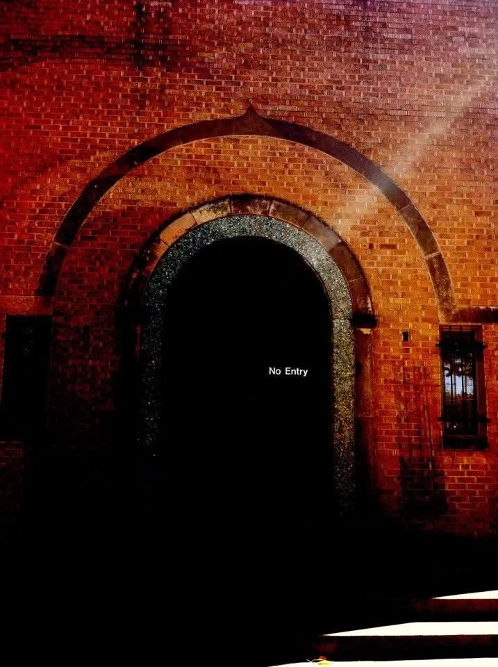 Dark arched doorway set in a red brick wall with a small window to the right, illuminated by warm lighting, and a sign that reads 'No Entry'