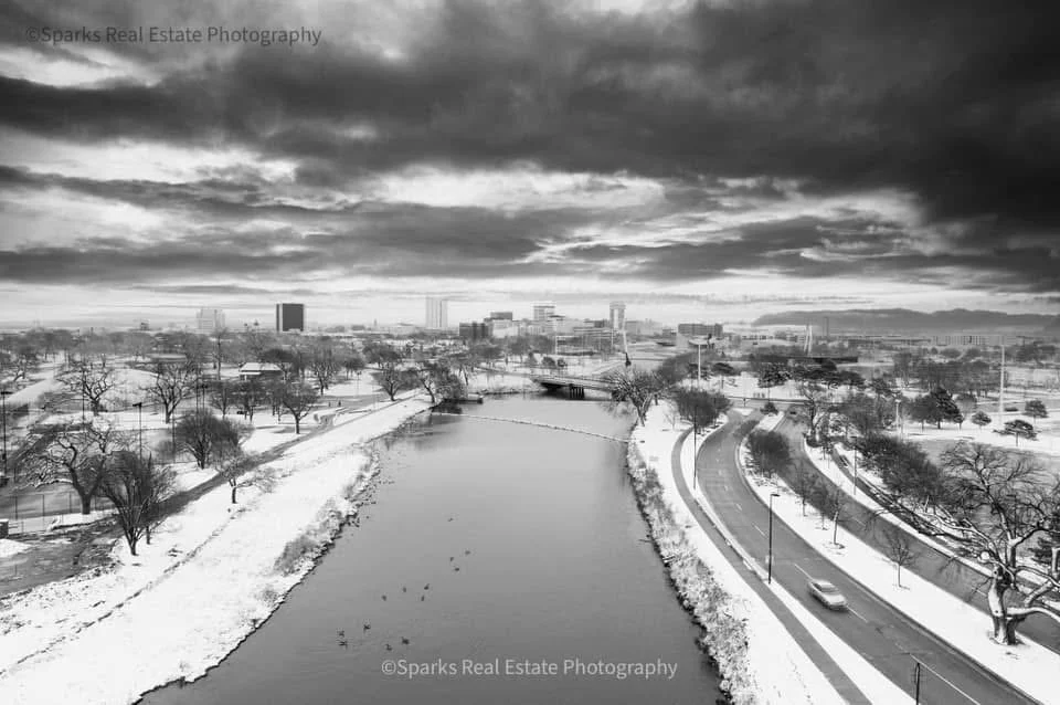 A black and white photo of a cityscape during winter showing a river, snow-covered trees, roads, and buildings under a cloudy sky.