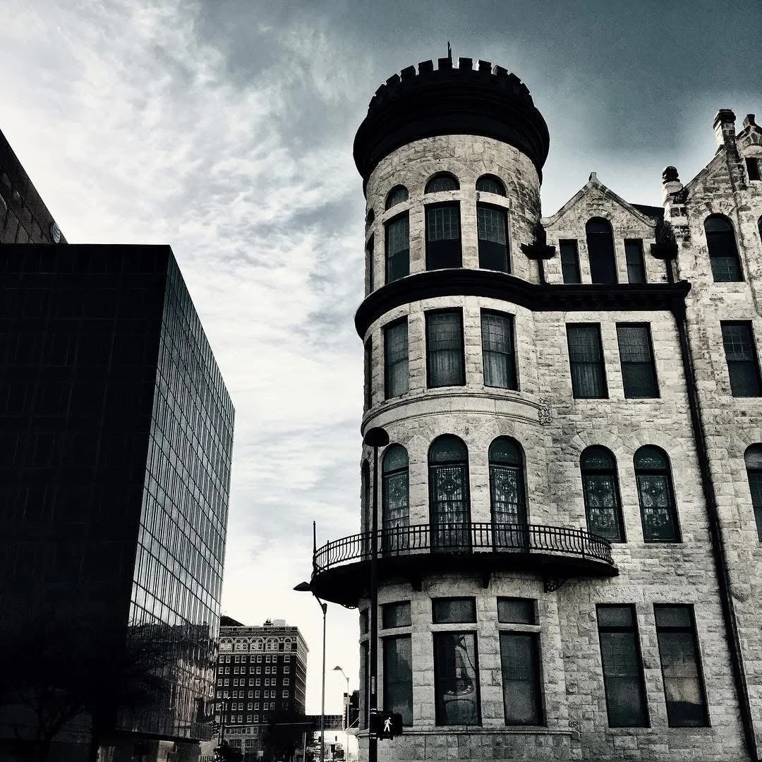 A historic stone building with turret and arched windows, contrasting with a modern black glass building nearby, under a cloudy sky.
