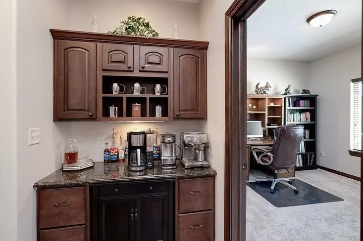 Home office with bookshelf, desk, and window, connected to a coffee station area with cabinets, coffee makers, and supplies.