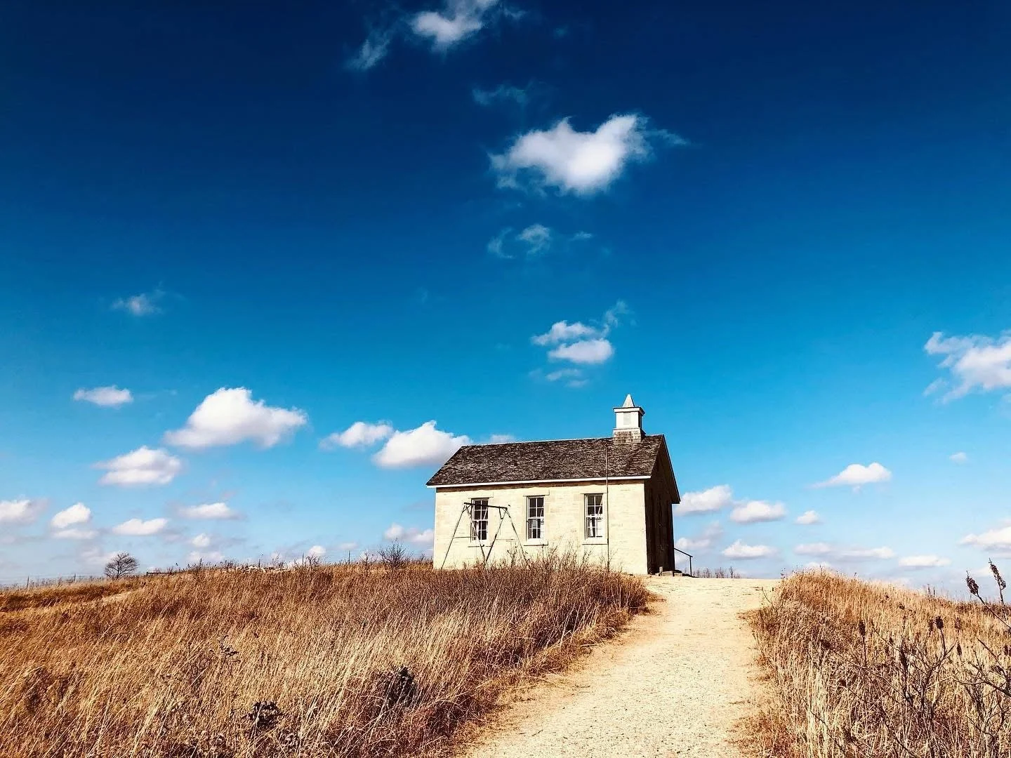 A small white house with a brown roof and a chimney, situated on a hilltop, surrounded by dry grass and a dirt path leading up to it under a bright blue sky with scattered white clouds.