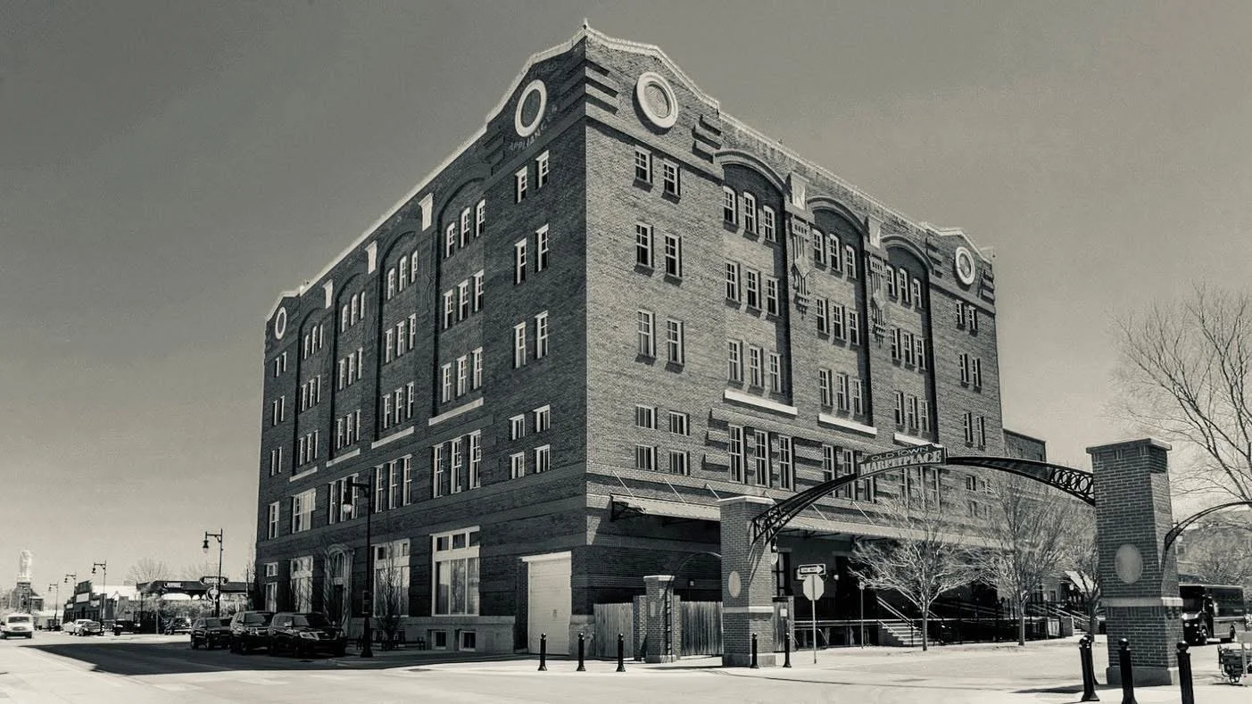 A large brick building with multiple floors, located at the corner of a street, with an arched entrance and trees in front.
