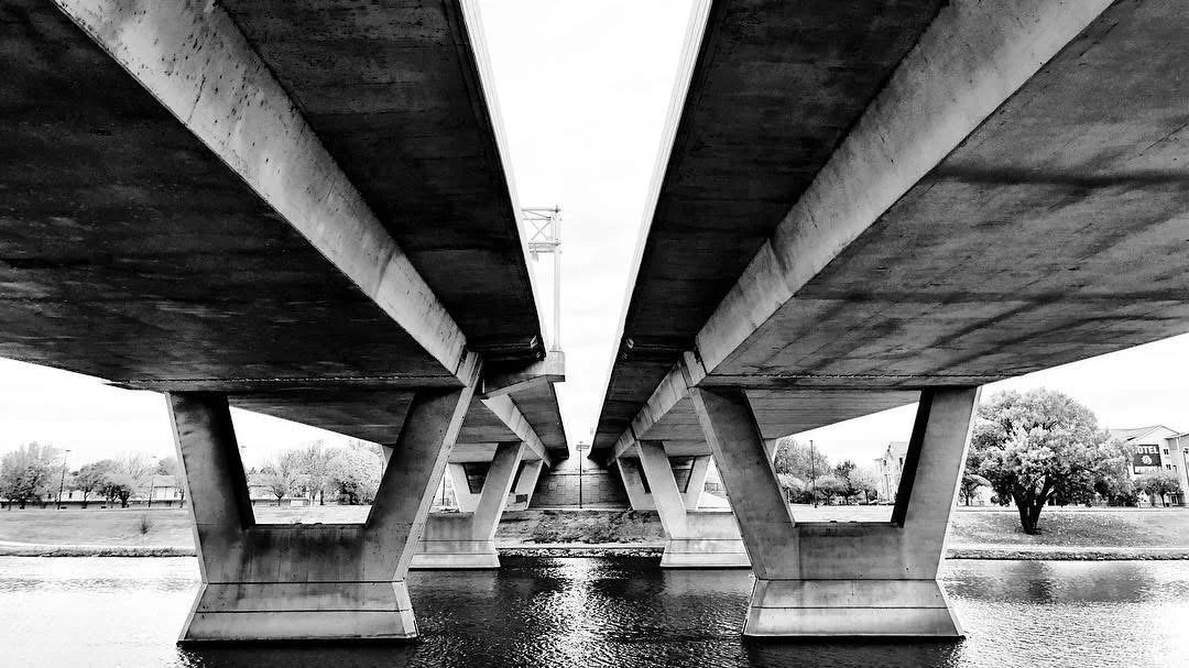 Black and white photo of a bridge with concrete supports spanning a body of water, taken from underneath looking up.