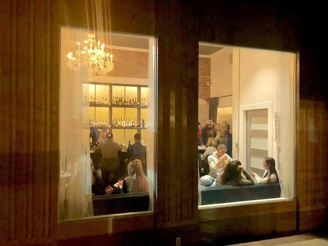 People sitting inside a bar or restaurant at night, with a large window showing the interior, including a chandelier and shelves with bottles behind the bar.