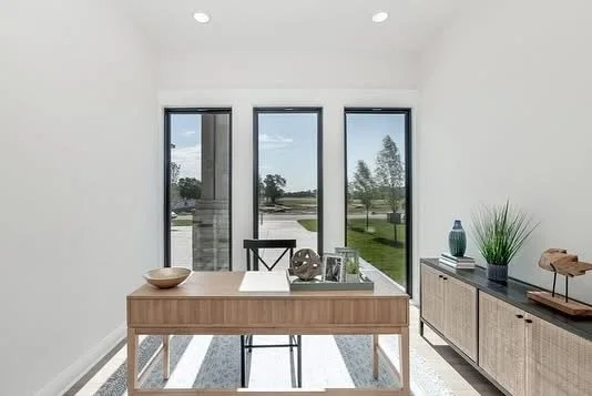 Minimalist dining room with a wooden table, black chair, and sideboard decorated with vases and artwork, with large windows overlooking a green outdoor area.