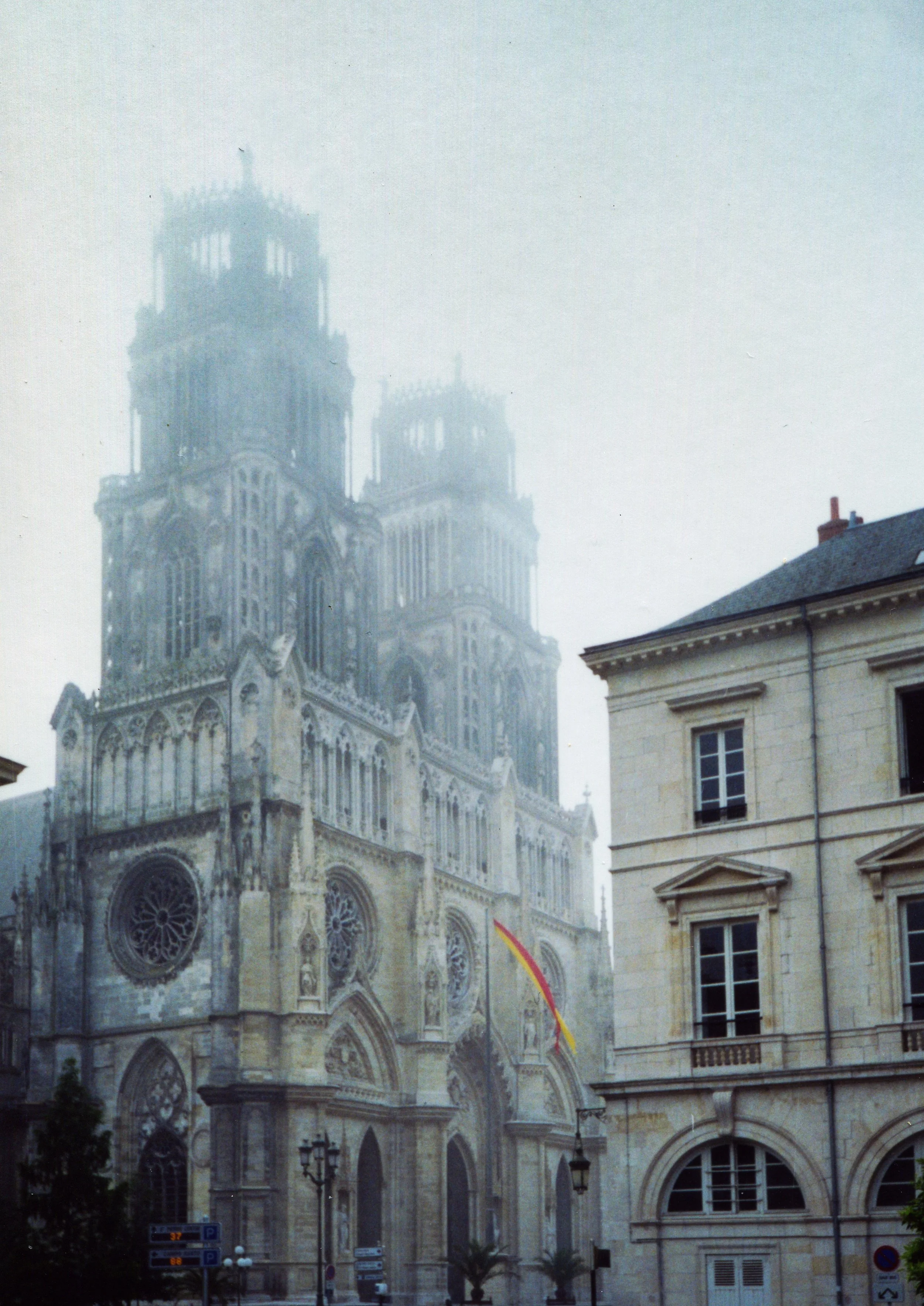A foggy view of the Gothic-style Basilica of Saint-Denis in France, with two tall towers, ornate architectural details, and a Spanish flag hanging outside.