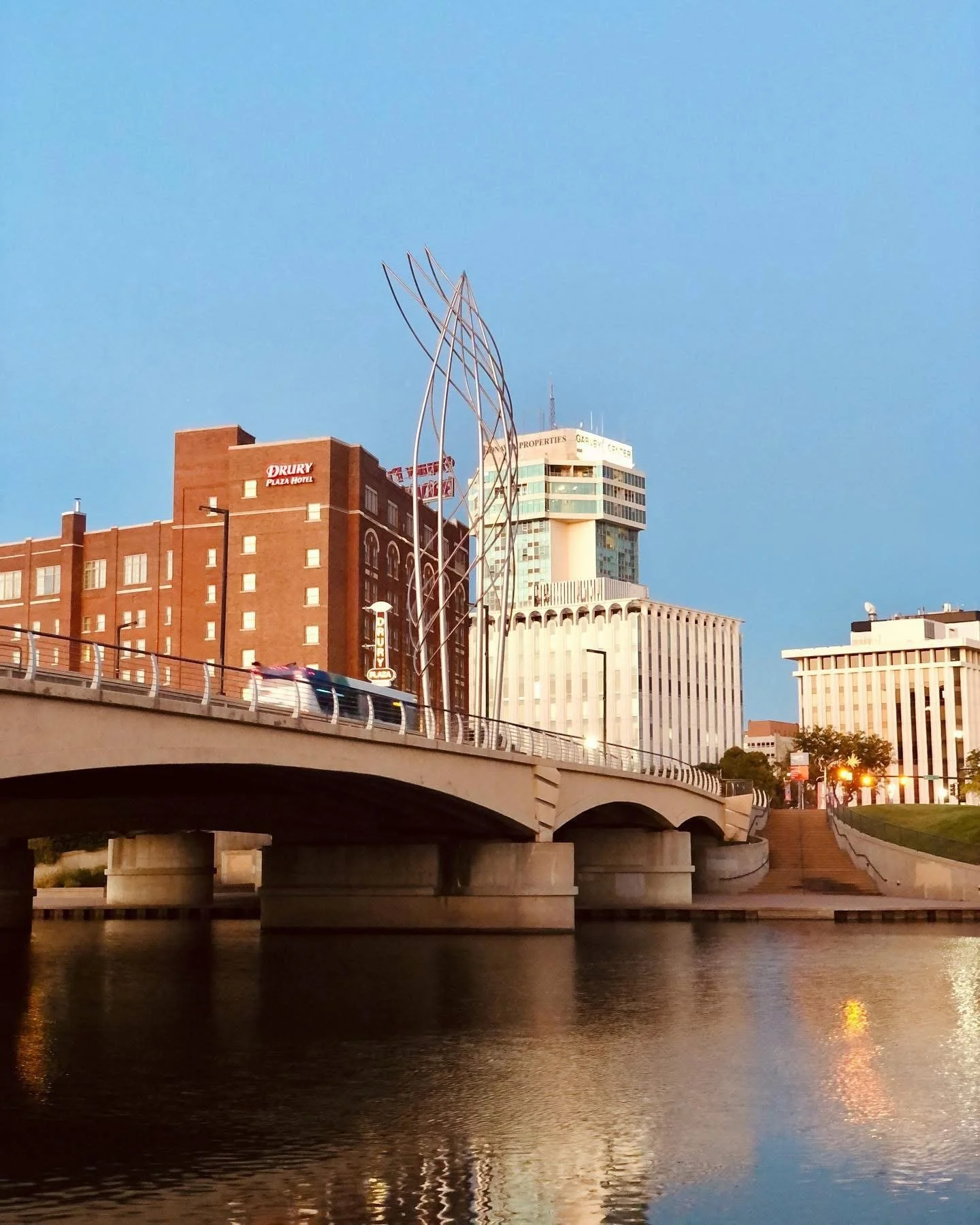 Cityscape with buildings, bridge over water, and a modern metal sculpture against a clear blue sky.