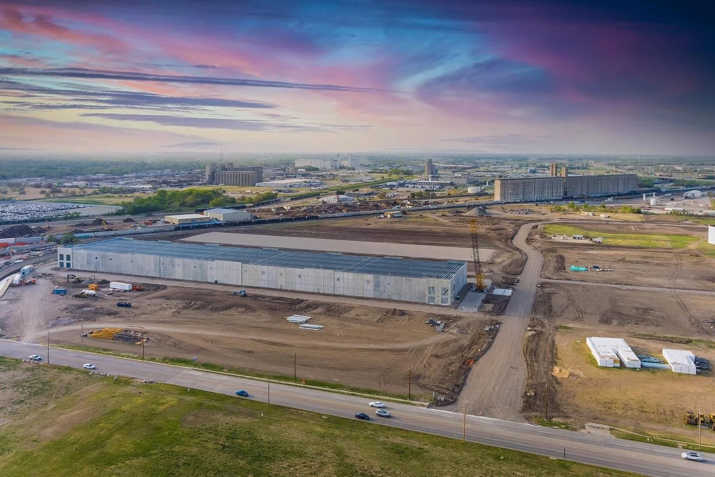Aerial view of a construction site with a large industrial building under construction, surrounded by dirt roads and equipment, with a city skyline and colorful sunset sky in the background.
