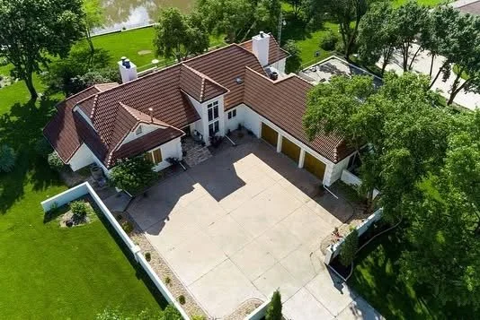 Aerial view of a large house with a brown tiled roof, white exterior, connected garage, and surrounding green lawn and trees.