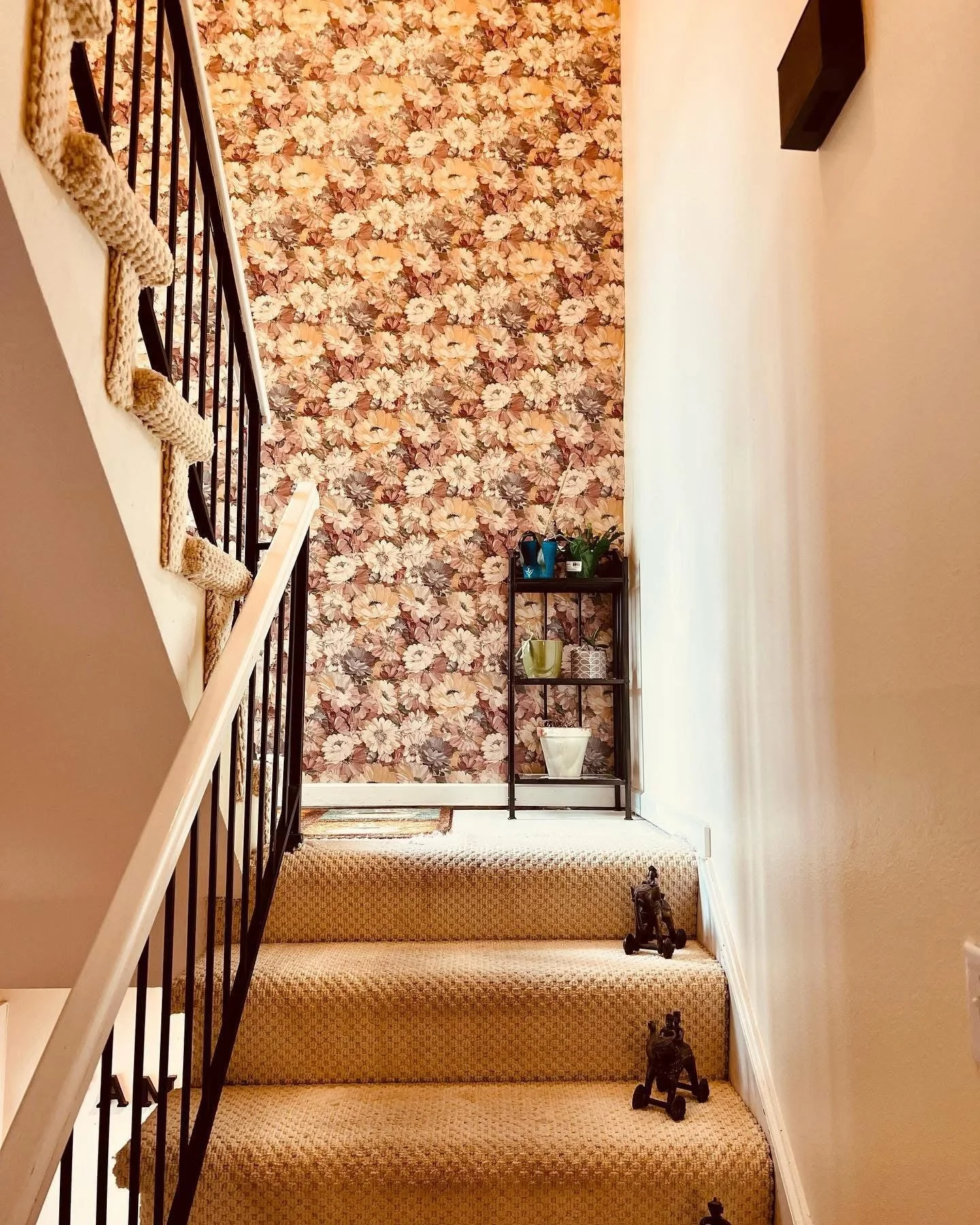 Interior view of a staircase with floral wallpaper at the top, beige carpeted stairs, black sculptures of animals placed on the steps, and a black metal shelf with potted plants at the top of the stairs.