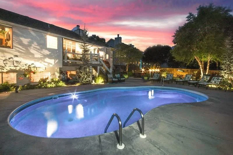 Residential backyard with a lit swimming pool, outdoor lounge chairs, trees, and a house with a balcony at sunset.