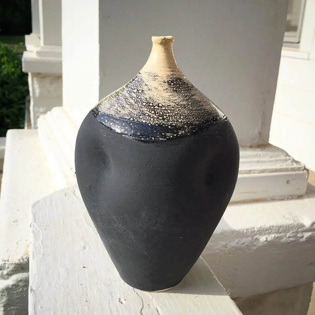 Black pottery vase with a textured, cream-colored top and crackled pattern near the rim, sitting on a white surface outside a building.