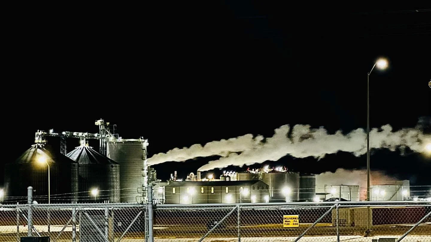 Night photograph of an industrial facility with silos and smoke stacks emitting white smoke, surrounded by a chain-link fence with yellow signs and illuminated by streetlights.