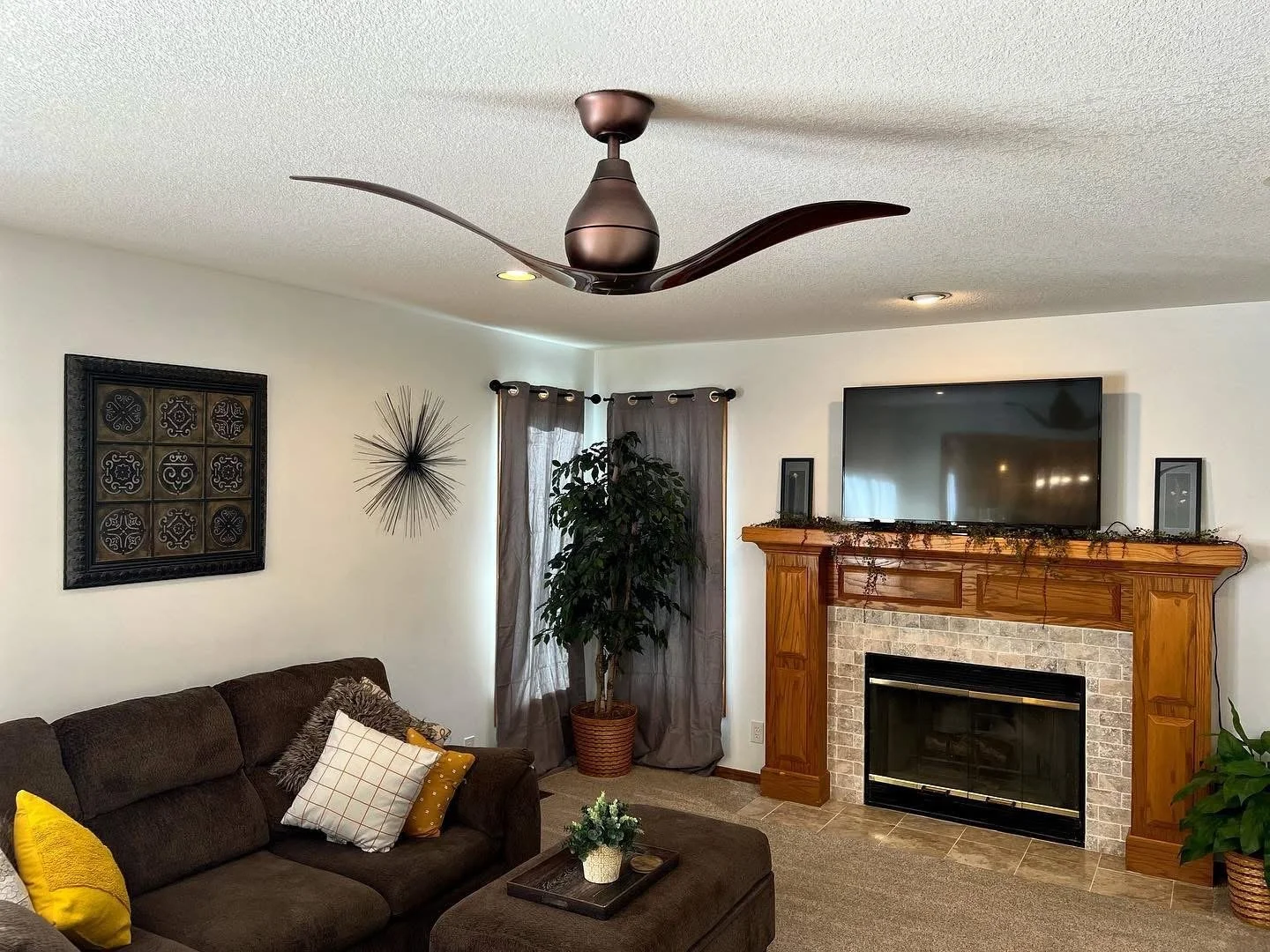 Living room with dark brown sofa, patterned pillows, a coffee table with a small plant, a wood fireplace with a TV above, decorative wall art, window with dark curtains, potted plants, a ceiling fan, and neutral-colored walls.